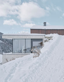 ​In einem kleinen Weiler der Tiroler Gemeinde Haiming, umgeben von ländlicher Idylle, erhebt sich ein neues Wohnhaus das nicht nur die Vergangenheit respektiert, sondern auch die Zukunft gestaltet. Sensibel fügt sich das neue Gebäude in die bestehende Umgebung ein und überrascht mit seiner differenzierten Baukörperplastik, die bei genauer Betrachtung eine faszinierende Geschichte erzählt. Bereits bei der Zufahrt zum Gebäude wird der Besucher von einer harmonischen Symbiose aus Vergangenheit und Gegenwart begrüßt. Das Eingangsgebäude mit Satteldach und brauner Holzfassade auf Straßenniveau zollen dem gegenüberliegenden Bauernhaus Tribut.  Über dieses gelangt man in den Wohnbereich welcher sich durch eine raumhohe Verglasung öffnet und einen Panoramablick auf die naturbelassene Umgebung bietet. Unterhalb der dieses Wohnbereiches finden die Pferde der hauseigenen Zucht ihr neues Zuhause. Die Betonfassade mit Bretterschalung verleiht dem unteren Teil des Gebäudes eine zeitgemäße Ästhetik, die sich wohltuend von der traditionellen Umgebung abhebt. Im Inneren kontrastieren die klaren Sichtbetonwände mit heimischen Eichenholz und naturbelassenen Stahl.   Wohnhaus LFD | Marlstein | ROECK Architekten | Innsbruck | Haus modern | Architekt Innsbruck | Sichtbeton | Wohnhaus Tirol | Stallgebäude | Pferdezucht | ROECK Architekten | Röck | Karlheinz Röck | Architekturbüro Röck