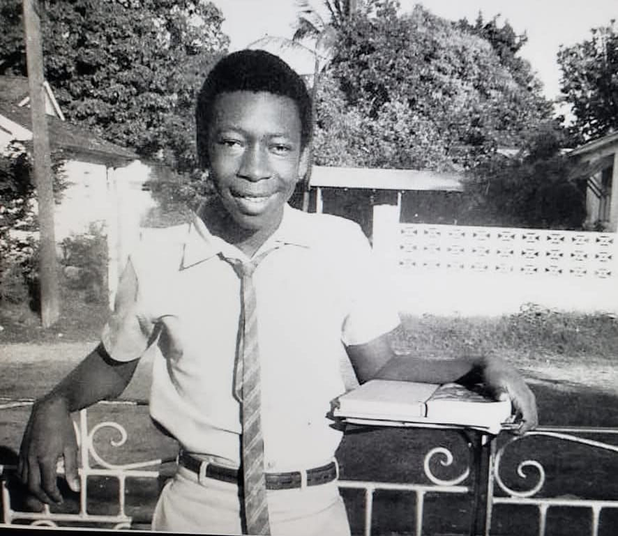 A young Dave Rodney, dressed in school uniform and holding books, leans on a fence. Trees and a house are visible in the background