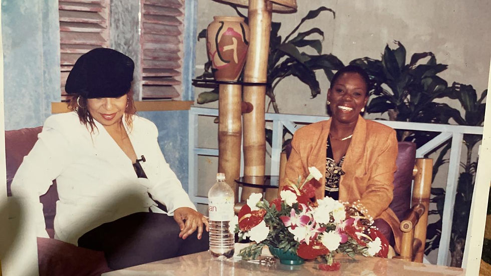 A smiling Darcy Tulloch, dressed in a yellow blazer, sits at a table with Roberta Flack on a light blue painted verandah. 