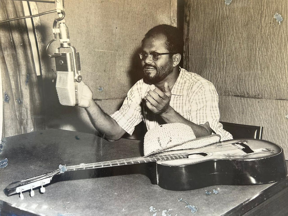 Monochrome photo of Carol Reckord seated in a recording studio, in front of a microphone. A guitar and straw hat rest on the table.