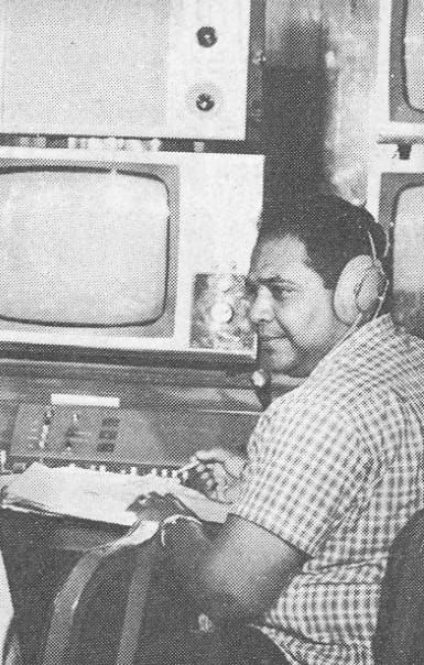 Mervyn Carby, clad in a checkered shirt and wearing headphones, operates equipment in a room with multiple monitors.