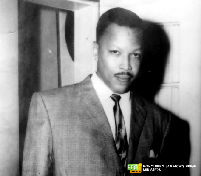 Monochrome upper-body portrait of Hugh Shearer in a suit and two-tone tie, standing indoors with a confident expression.