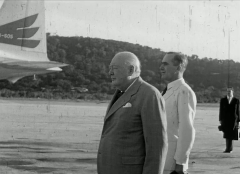 Monochrome image of Winston Churchill in a suit on an airstrip, with two men and the tail of a plane visible against forested hills.