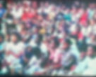 A large group of children sit closely together in a studio, appearing attentive. They wear colorful clothing with various patterns.