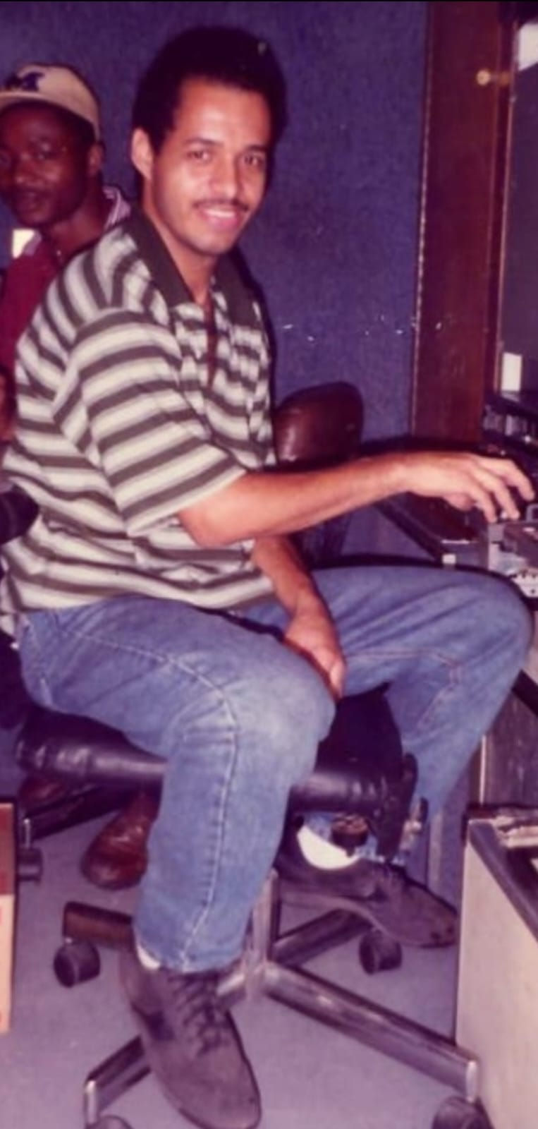Neville McKenzie, smiling in a striped shirt, sits at a control panel. A man in a yellow hat is partially visible in the background.