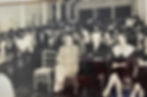 Black and white photo of a crowded auditorium. People sit and listen attentively, dressed in formal attire