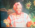'Miss Lou,' dressed in a traditional bandana outfit and headscarf, gestures expressively in front of a floral backdrop.