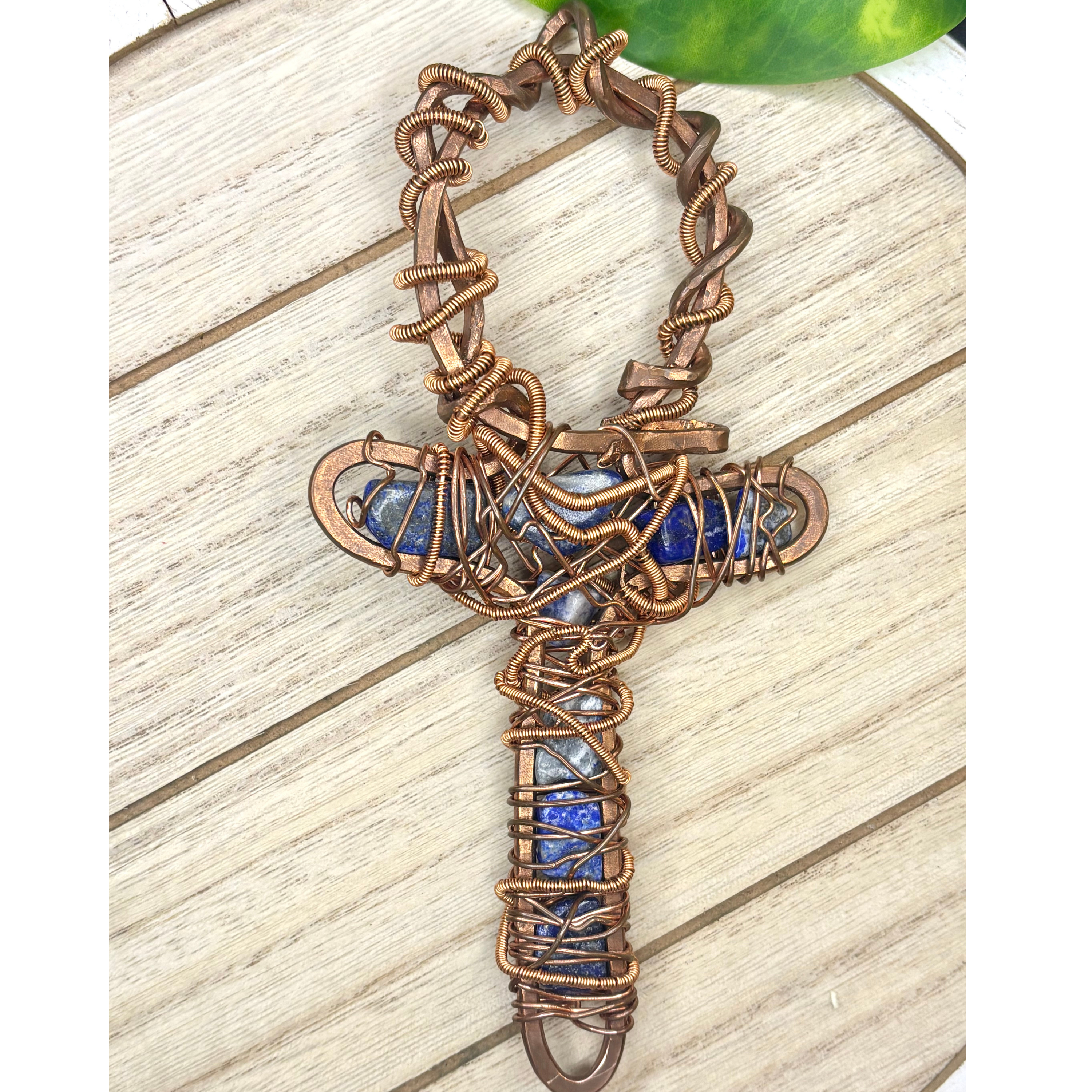 Handmade copper wire wrapped lapis lazuli ankh altar decor displayed flat on a white wood surface with a green houseplant in