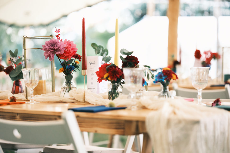 Ein Blick auf einen festlich gedeckten Holztisch unter einem Zelt, dekoriert im "Festival-Colors of Love"-Stil. Die Tischdekoration umfasst leuchtende Blumenarrangements in Pink, Rot, Orange und Blau in kleinen Glasvasen, hohe rote und gelbe Kerzen, einen cremefarbenen Musselin-Tischläufer und klare Weingläser. Blaue Servietten setzen farbige Akzente. Die Szene ist hell, farbenfroh und einladend für eine besondere Feier unter dem Stretchtent ein.