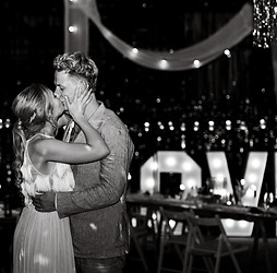 Euer romantischer Hochzeitsmoment: Ein frisch vermähltes Paar teilt einen Kuss bei eurer Hochzeitsfeier. Im Hintergrund sorgen funkelnde Lichterketten und beleuchtete "LOVE"-Schild für eine magische und unvergessliche Atmosphäre, ganz nach dem Motto "Love is in the air".