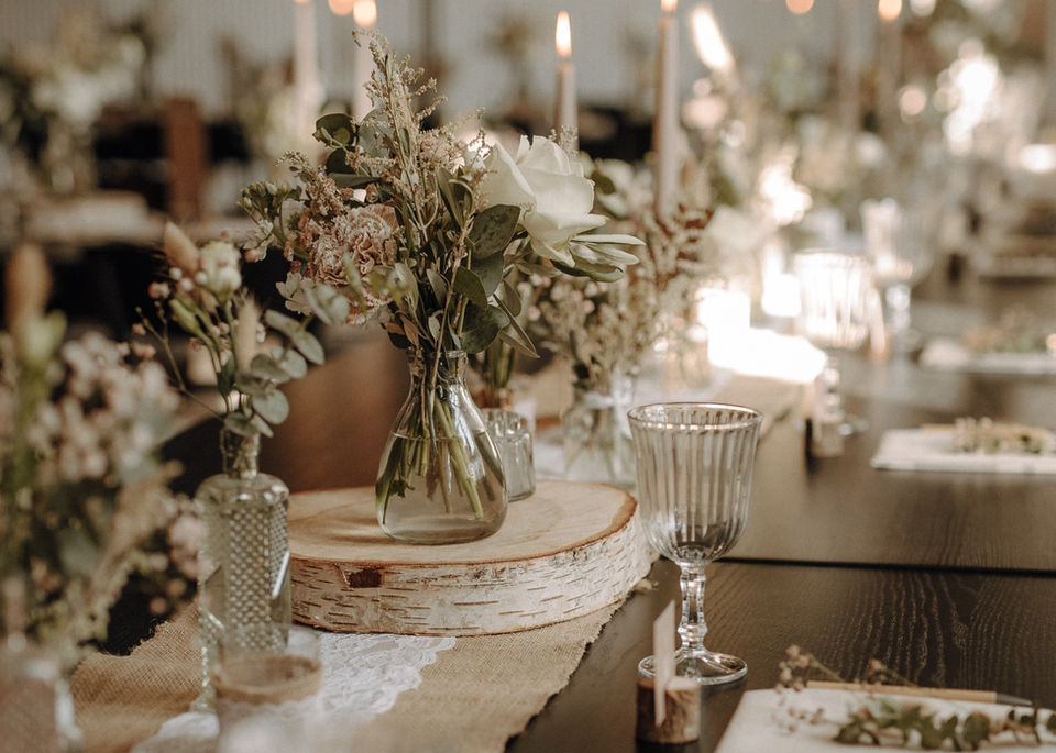 Stellt euch eure Tischdekoration im Vintage-Bohostil auf einem dunklen Holztisch vor. Die Dekoration umfasst zarte Blumenarrangements mit natürlichen Blüten und Eukalyptus in Glasvasen, hohe Kerzen auf Holzscheiben, einen beigen Jute-Tischläufer mit weißer Spitze und filigrane Weingläser. Die Szene ist stimmungsvoll beleuchtet und strahlt eine romantische, natürliche Atmosphäre aus, ideal für eine Hochzeitsfeier.