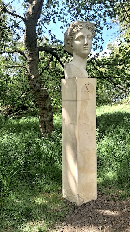 A bust sits atop a column fashioned in stone, set within a pleasant wooded location with the sun shining through the trees.