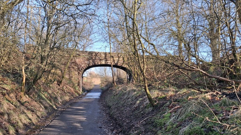 Stone bridge over a narrow path surrounded by leafless trees and grassy banks on a sunny day, creating a peaceful and serene mood.
