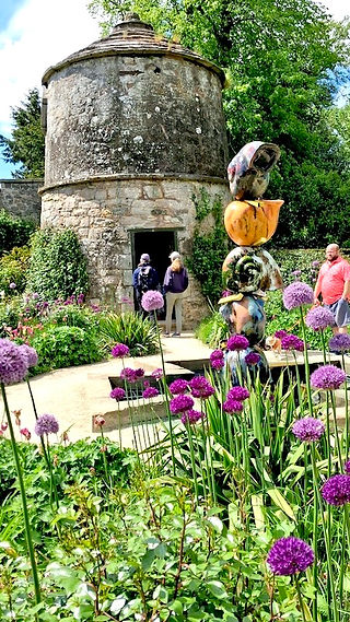 A brightly coloured sculpture foreground with alliums in full bloom on a sunny day. At the rear lies an ancient dovecotek