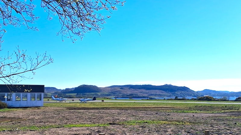 Small planes on a grassy airfield at Oban Airport, with a distant view of hills under a clear blue sky. A tree with bare branches frames the scene.
