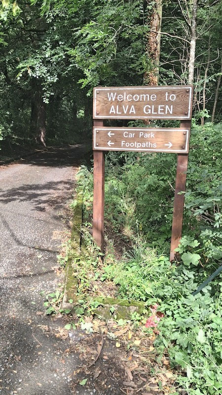 Wooden sign reading Welcome to ALVA GLEN with arrows to car park and footpaths beside a forest path.
