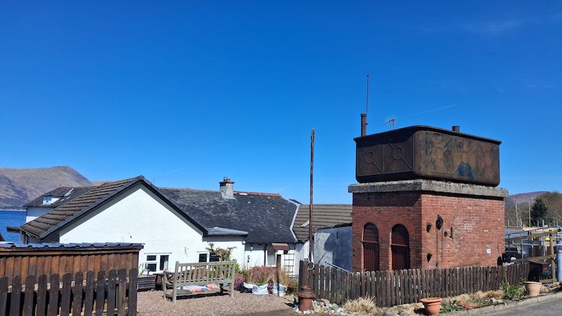 Brick building at Kintallen Station with a rusty metal tank on top, next to a white house with a brown roof. Clear blue sky and mountain in the background.