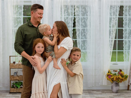 A heartwarming family moment at Honey B's Greenhouse in Howe, Texas, captured by Precious Gems Photography. A mother shares a tender kiss with her young son, surrounded by her partner and children, with light curtains, sunflowers, and floral art enhancing the cozy, welcoming atmosphere