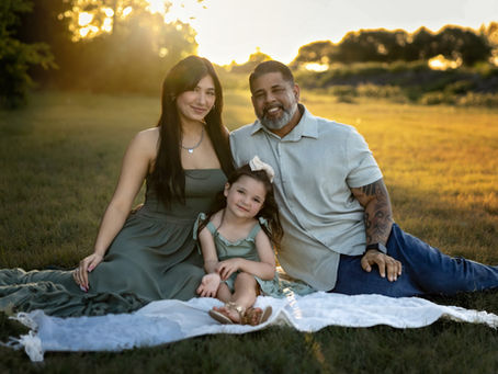 A family enjoys a sunny afternoon on a blanket in a picturesque field in Haslet, Texas, captured by Precious Gems Photography. The parents wear green and a light shirt, while their daughter dons a matching green dress.