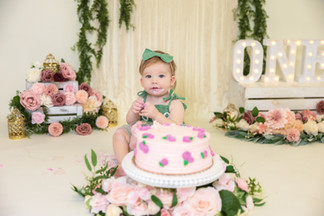 Baby with pink cake, wearing a green bow, amidst floral decor. "ONE" sign in the background, set in a festive, pastel-toned scene. Cake Smash Session done at Creative Chelle Studios in Saginaw Texas by Precious Gems Photography. 