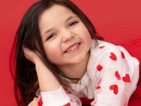 Smiling toddler girl lying on red studio backdrop wearing a white heart top and red skirt during Valentine mini session at Precious Gems Photography in Haslet Texas