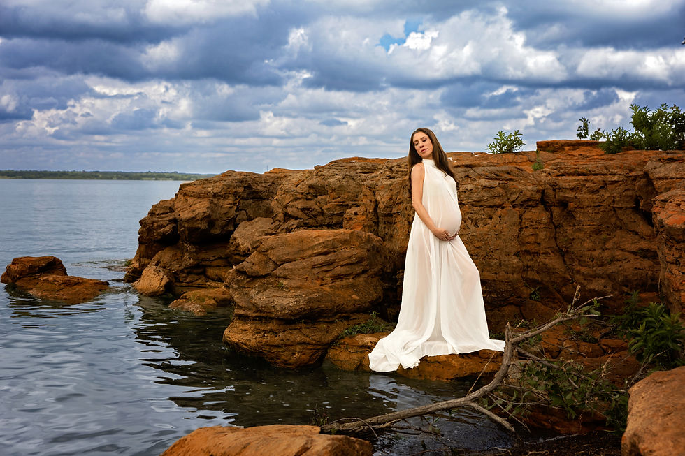 Pregnant woman in a flowing white dress stands on rocky lakeshore beneath dramatic cloudy sky.