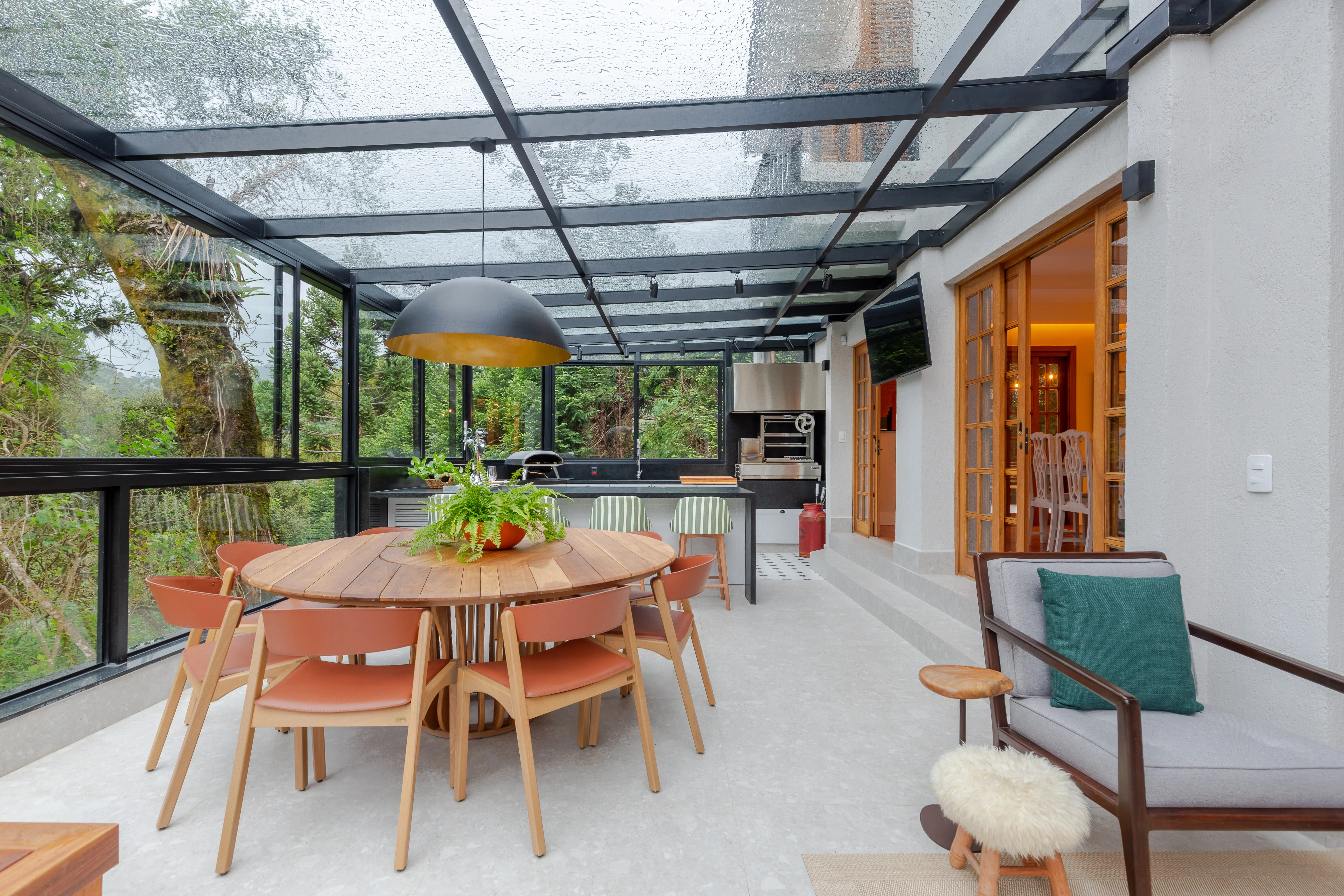Sala de jantar envidraçada com mesa redonda e cadeiras em tons quentes, cercada por vegetação e iluminada por pendente central.