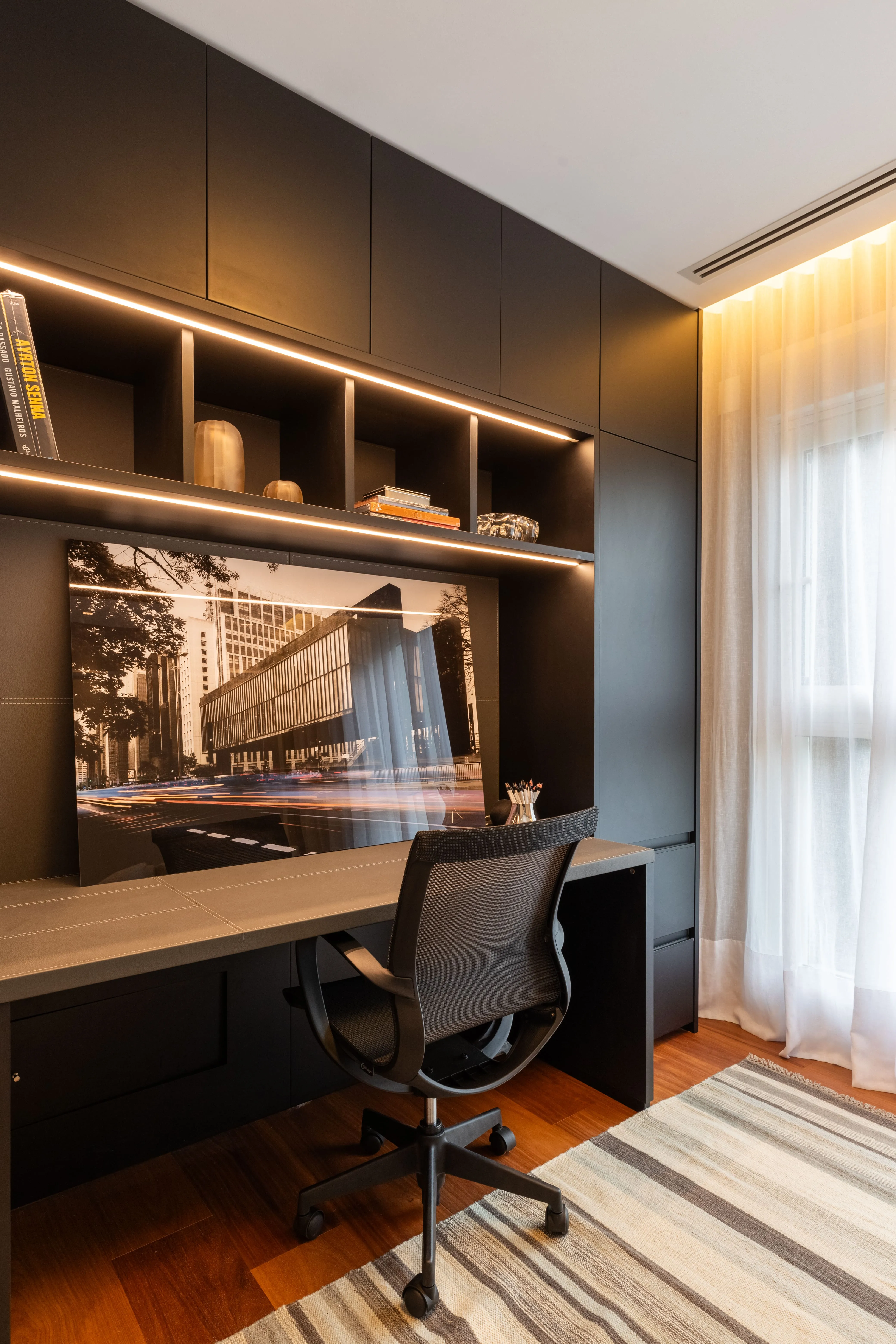 Escritório em casa com estante iluminada em marcenaria preta, mesa de trabalho e cadeira ergonômica próximo à janela com cortina leve.