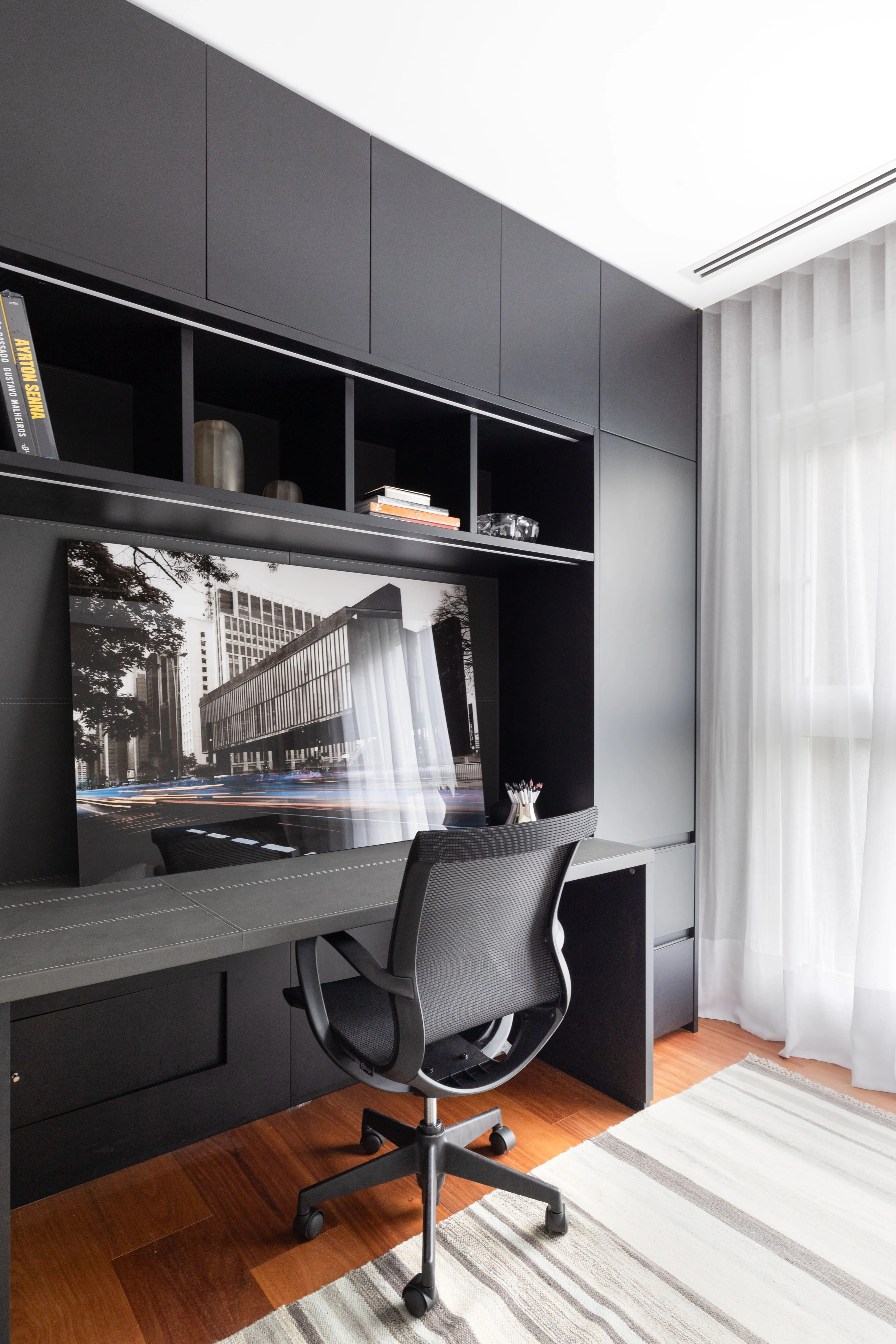 ome office com marcenaria preta planejada, mesa de trabalho, nichos com iluminação embutida e cadeira ergonômica ao lado de cortina translúcida.