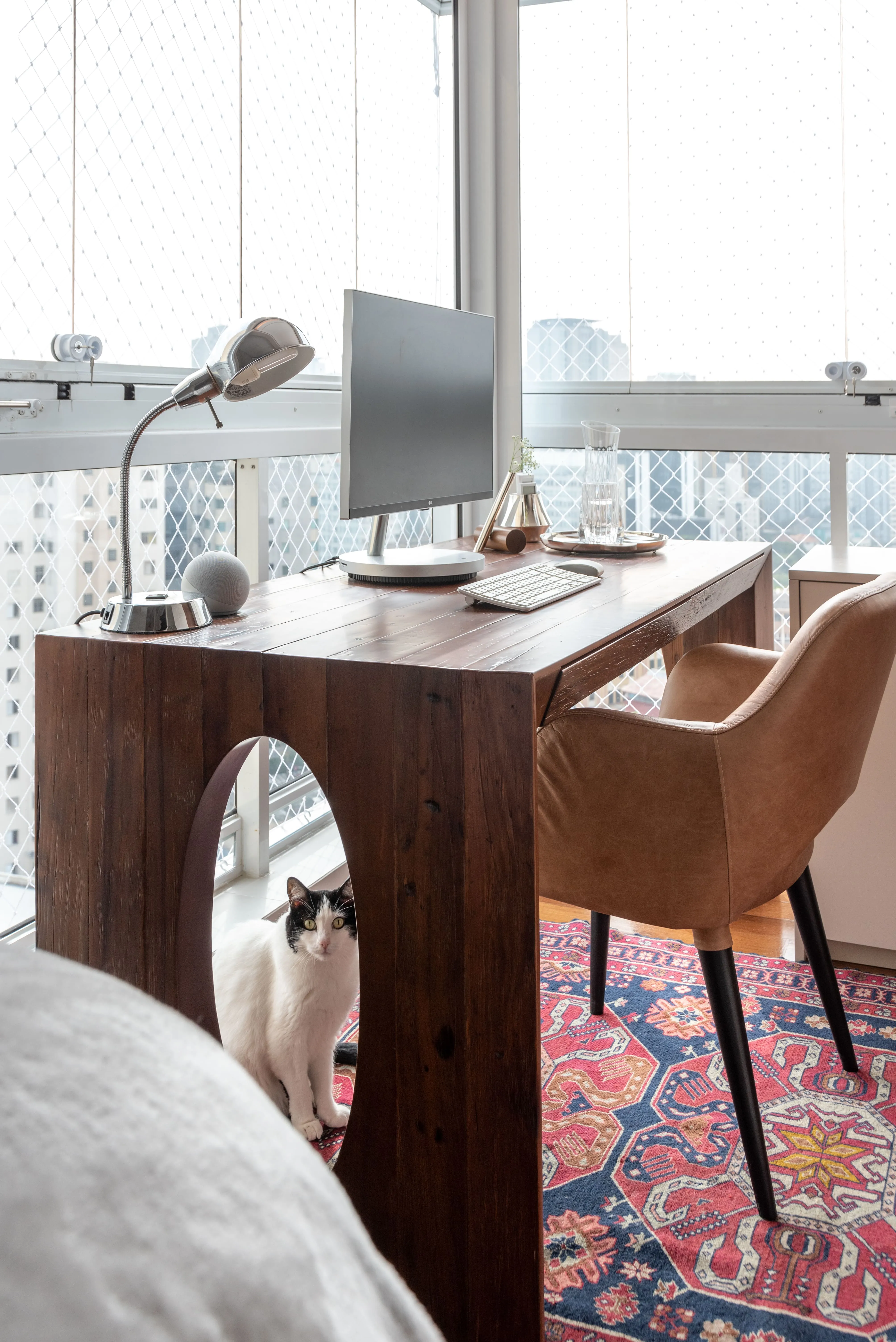 Home office compacto com mesa de madeira escura, cadeira estofada em couro e computador posicionado próximo à janela com vista urbana.