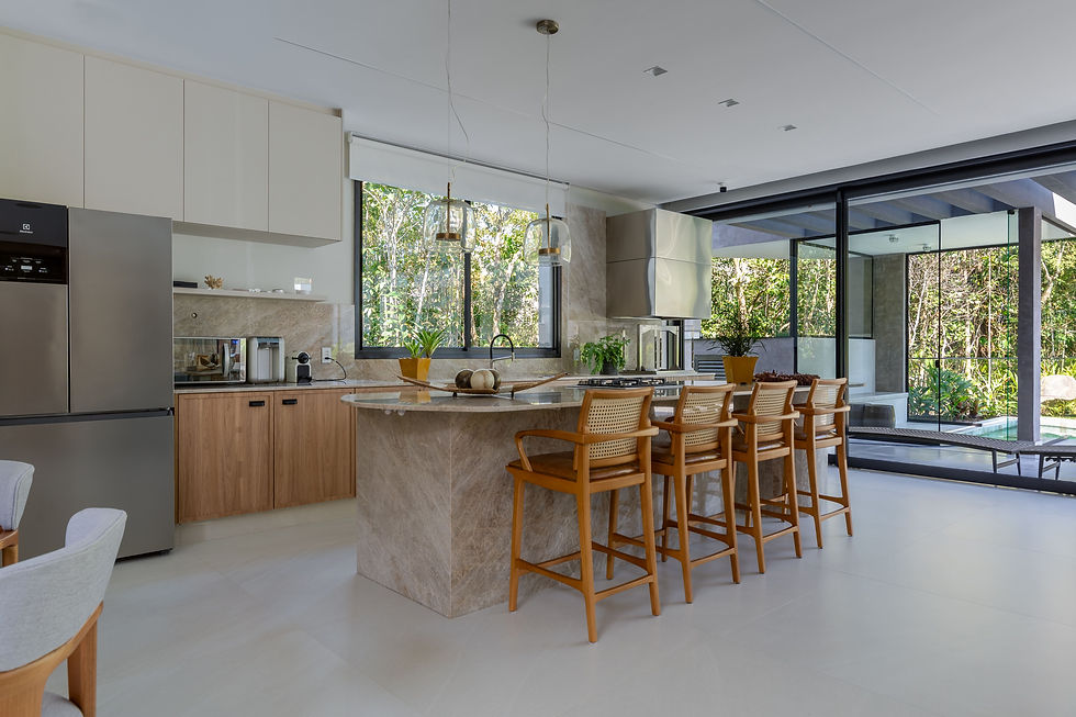 Cozinha moderna integrada com ilha central em pedra, armários claros e madeira, bancada com banquetas, eletrodomésticos embutidos e grandes portas de vidro que conectam o ambiente ao jardim externo.