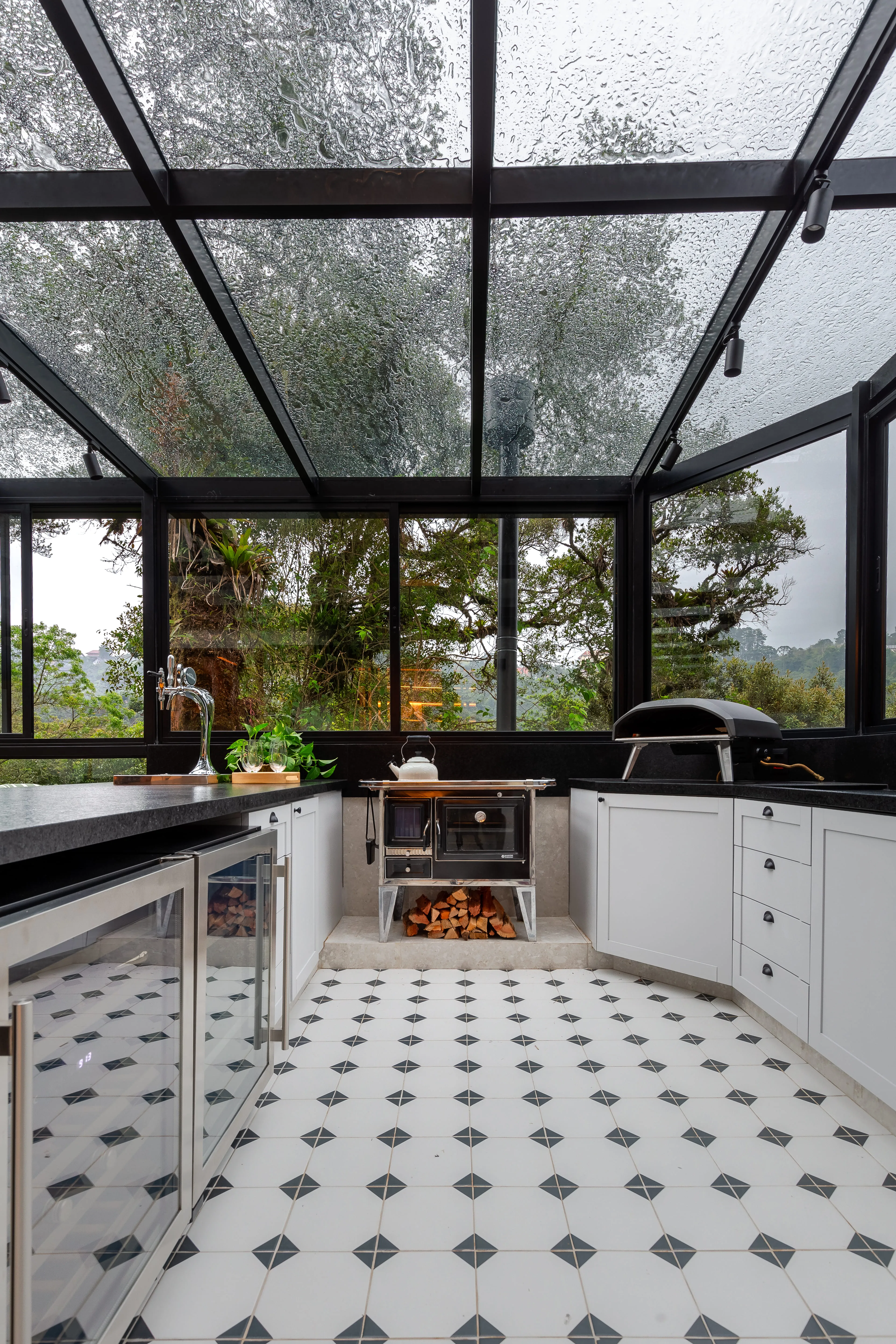 Cozinha com armários brancos, bancada em madeira e piso com padrão geométrico, sob teto envidraçado que permite entrada de luz natural.