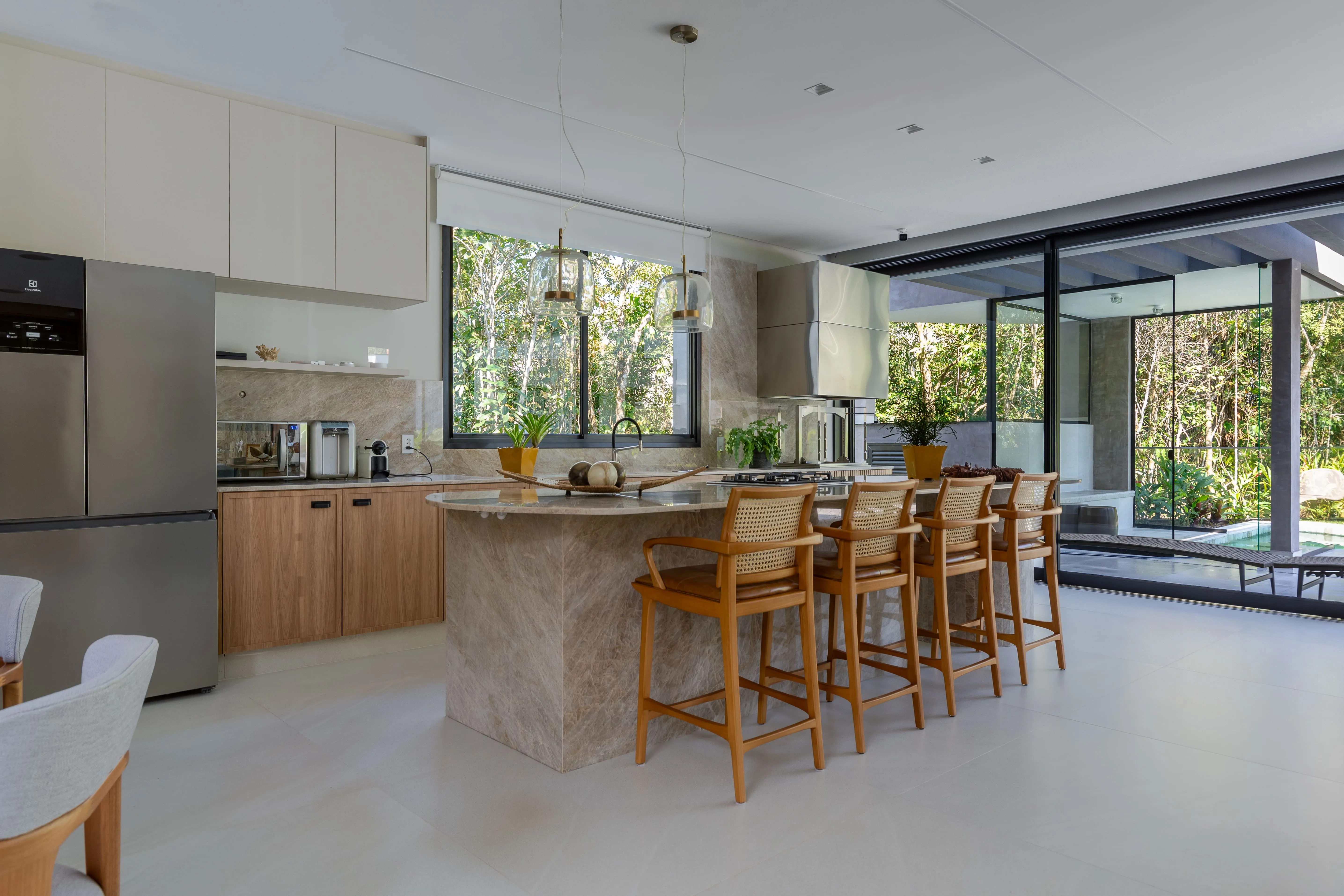 Cozinha integrada com ilha em pedra, banquetas de madeira e armários claros, iluminada por grandes aberturas com vista para o jardim.