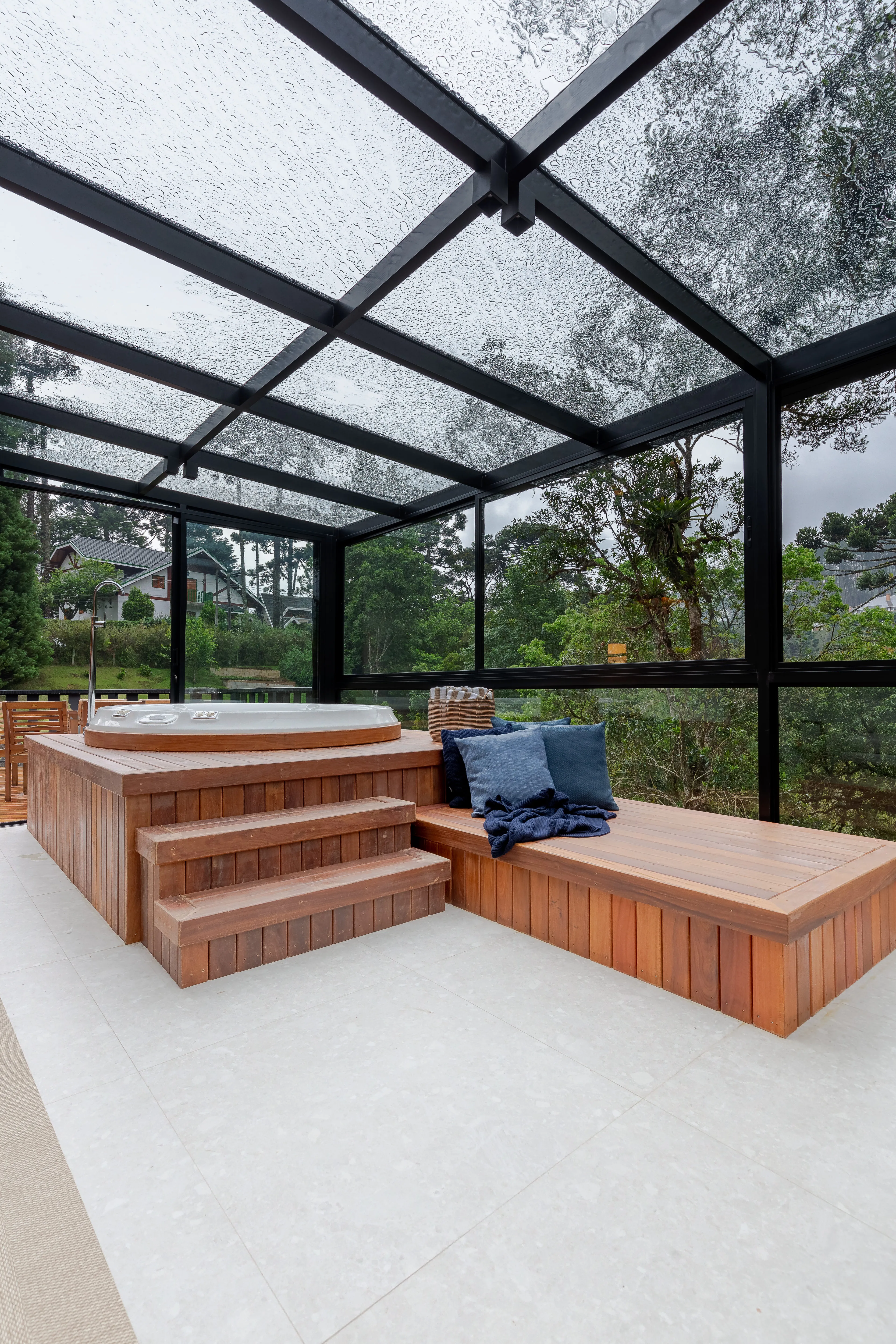 Espaço de convivência com deck de madeira em níveis, almofadas decorativas e fechamento em vidro, integrado à paisagem verde externa.