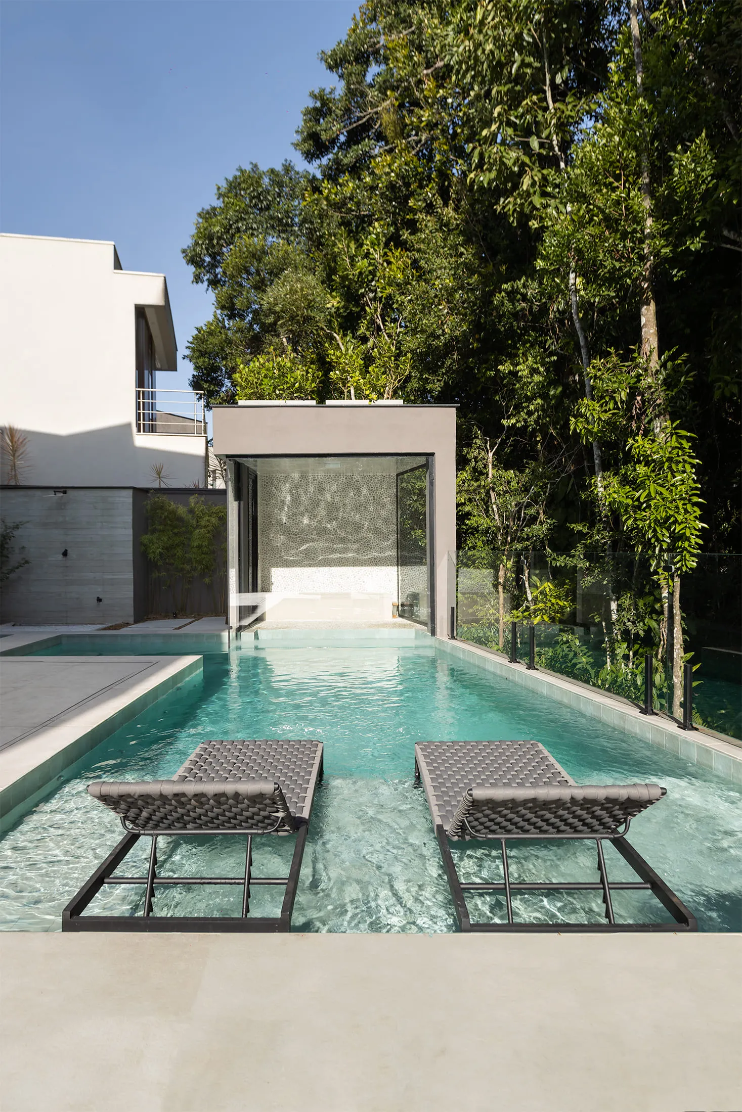 Piscina estreita e alongada cercada por vegetação, com espreguiçadeiras parcialmente submersas e arquitetura moderna ao fundo com portas de vidro.