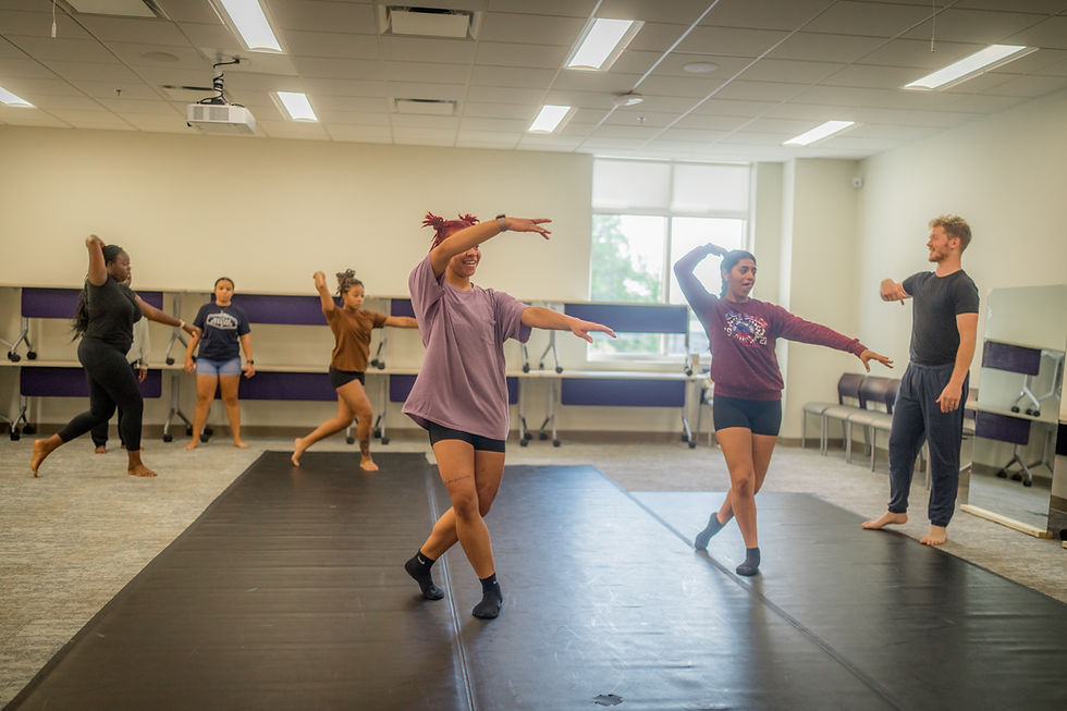 Five dancers attempt technical exercises across the floor while their dance teacher instructs from the front of the room.