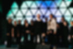 Young performers surround a soloist for the finale of a Christian musical song. All focus upward as green and blue colors light the stage.
