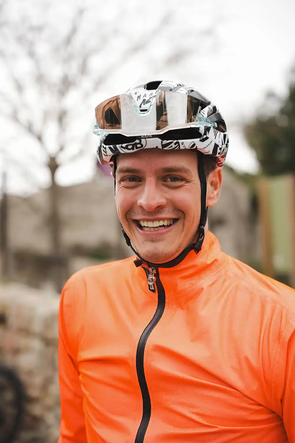 Portrait eines lachenden Rennradfahrers mit orangefarbener Windjacke und Helm im  808 Scyence Trainingcamp.