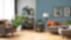 Modern living room with beige furniture, orange pillows, plants, and wall art. A blue accent wall and wooden floor create a cozy ambiance.