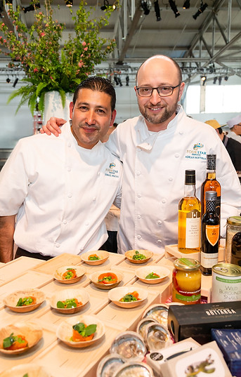 two chefs smiling warmly with scallop ceviche dishes and bottles of olive oil and provisions