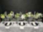 Un sofisticado diseño clásico de mesa de boda con una impactante paleta de colores en blanco y negro. Los centros de mesa consisten en elegantes arreglos florales blancos en jarrones de cristal transparente y elementos decorativos más oscuros, posiblemente negros. Entre las flores blancas más destacadas se encuentran el Lilium Roselily White, el delicado Crisantemo Báltico, el encantador Rosa Lady White, el ligero Phlox White y la delicada Gypsophila, acentuadas por la oscura Scabiosa con forma de botón. Velas blancas altas de diferentes alturas se intercalan por toda la mesa. La mesa está decorada con mantelería blanca impecable, elegantes platos y cubiertos negros, creando una estética elegante y atemporal.