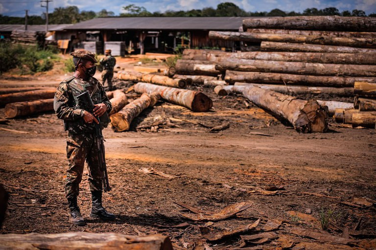 A foto mostra um homem usando uma farda do exército brasileiro, de máscara e segurando uma arma de fogo parado em pé. Atrás dele se vê troncos de árvores cortados e empilhados próximo a um galpão.