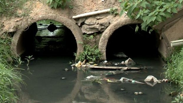 A foto mostra um córrego com dois bueiros cheios de água escura e lixo boiando.