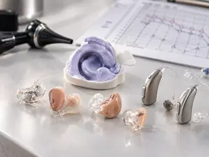 Audífonos sobre medida intracanal y retroauricular junto a un molde de impresión de oído y un otoscopio, con un audiograma al fondo, sobre una superficie clara bajo luz natural.