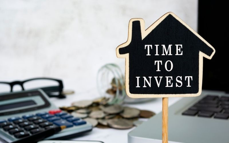 Miniature house-shaped sign reading “Time to Invest” with calculator, coins and laptop in background—symbolising financial planning.