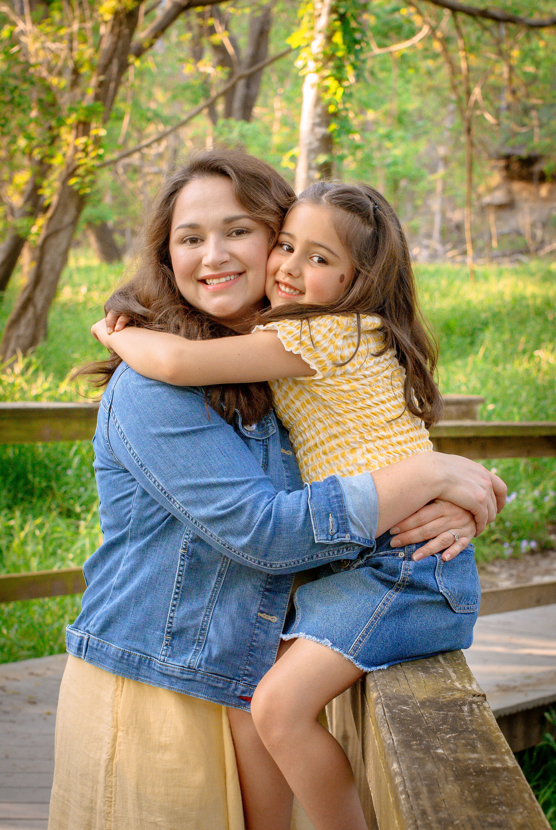 A mom and daughter embrace wearing yellow and blue. They have green trees behind them.