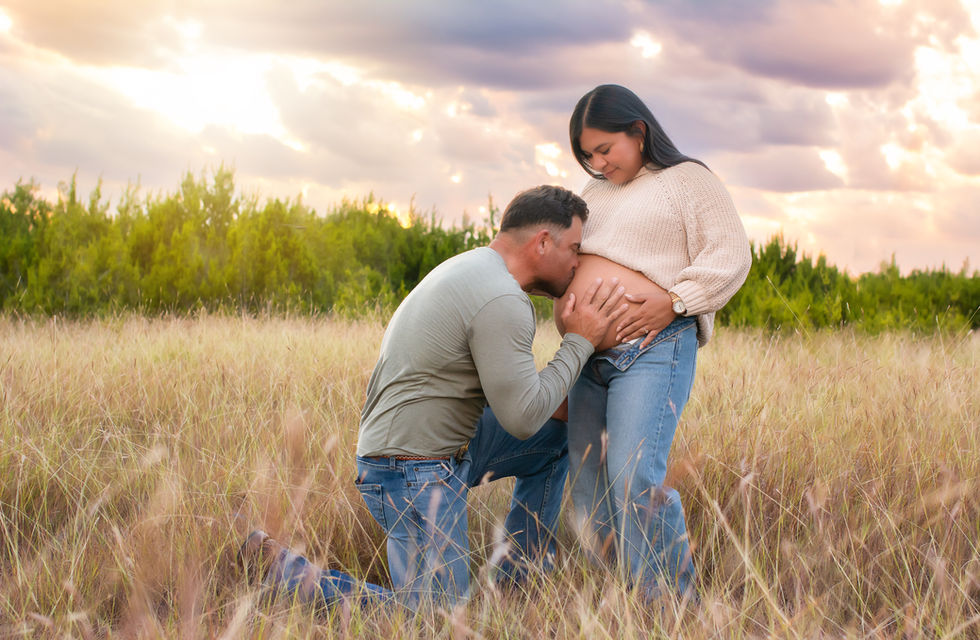 A husband kisses the wife's pregnant belly in a field in front of a sunset.