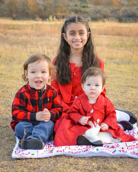 Three children wearing Christmas attire sit on a blanket in front of a lake in Cedar Park, Texas. Megumi Rooze is an affordable photographer in Cedar Park offering mini sessions for $100.