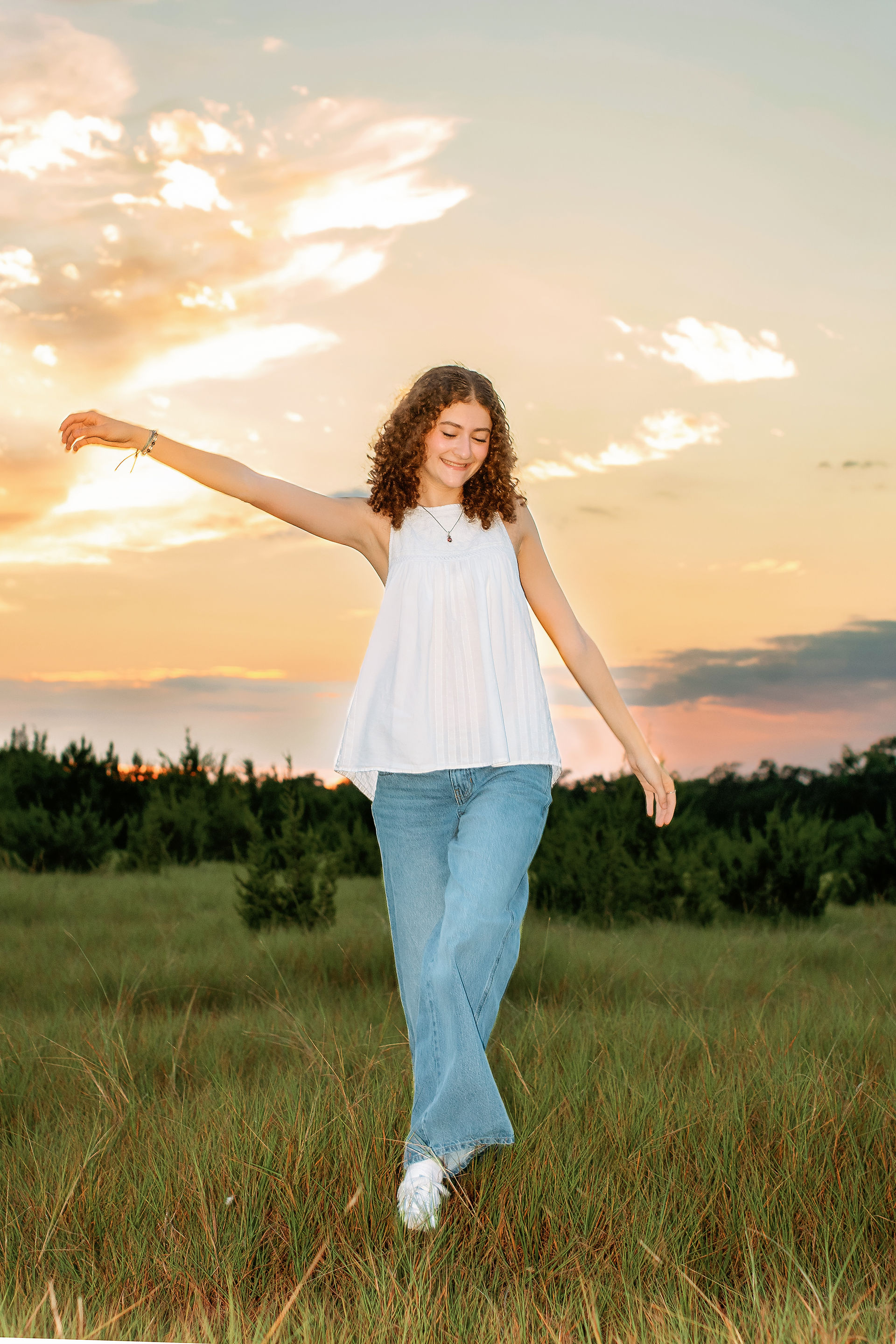 A Senior Girl in a white shirt and jeans walk through some grass with a Sunset behind her.