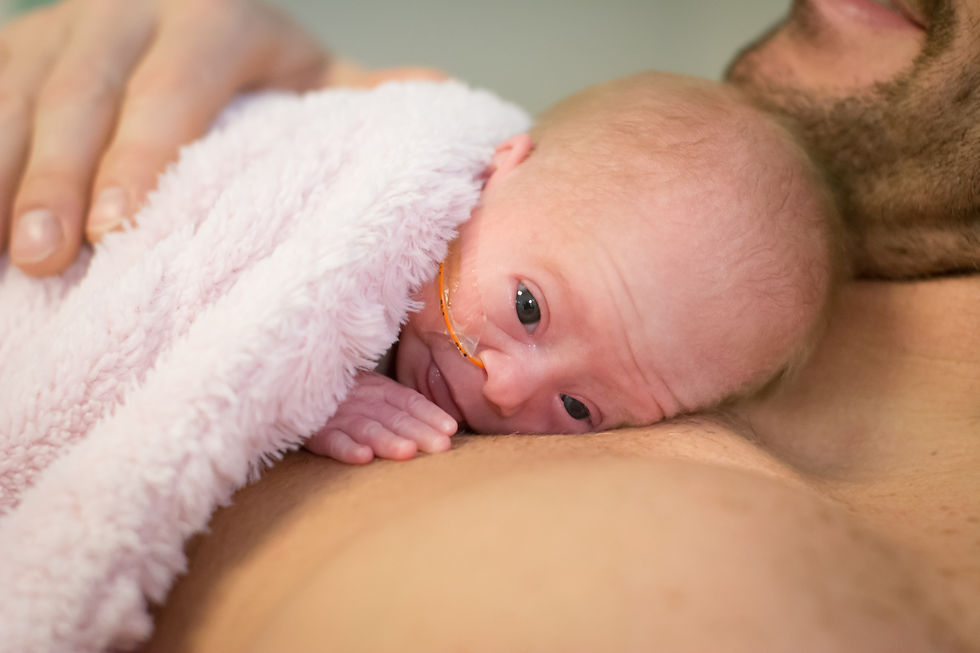 Newborn snuggles skin-to-skin on an adult's chest, wrapped in a pink blanket. Tube seen on baby, atmosphere is warm and nurturing.